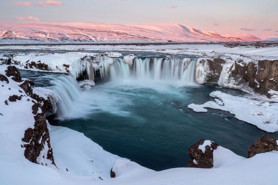 Una vasta cascata con i bordi ghiacciati si riversa in una piscina turchese in un paesaggio vasto e innevato al tramonto.
