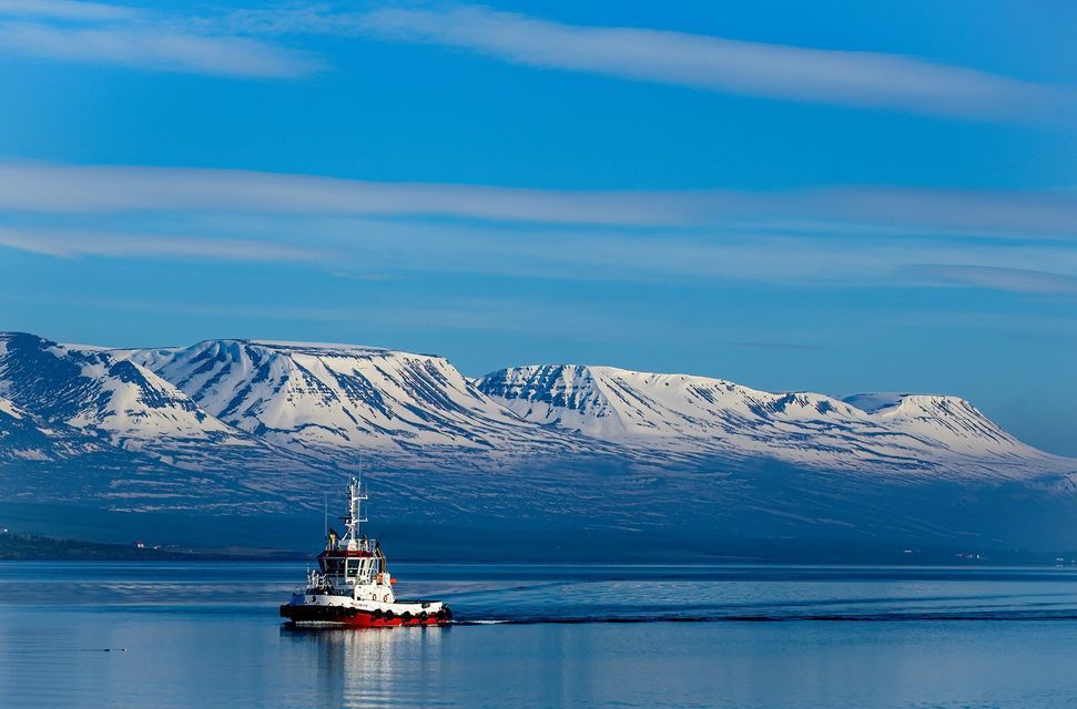 Un rimorchiatore rosso e bianco naviga su acque calme e blu, con un'ampia catena montuosa innevata sullo sfondo sotto un cielo sereno.