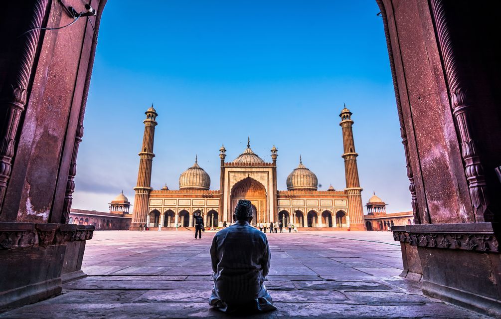 Un uomo visto di spalle si inginocchia all'interno di un arco di pietra, guardando verso una grande moschea con minareti al di là di un cortile.
