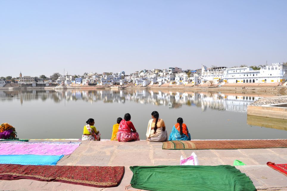 Cinque donne in sari colorati siedono su gradini di pietra in riva a un lago, con tessuti ad asciugare in primo piano e una città riflessa nell'acqua.