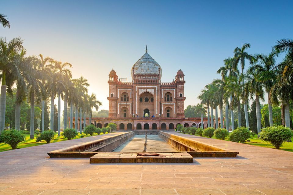 A red sandstone mausoleum with a large dome at the end of a long reflecting pool, flanked by palm trees under a clear sky.