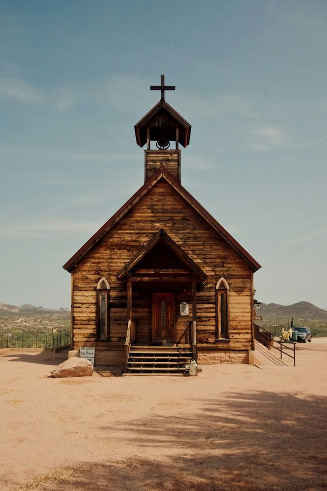 Una chiesa rustica in legno con campanile si erge in un paesaggio desertico sotto un cielo limpido.