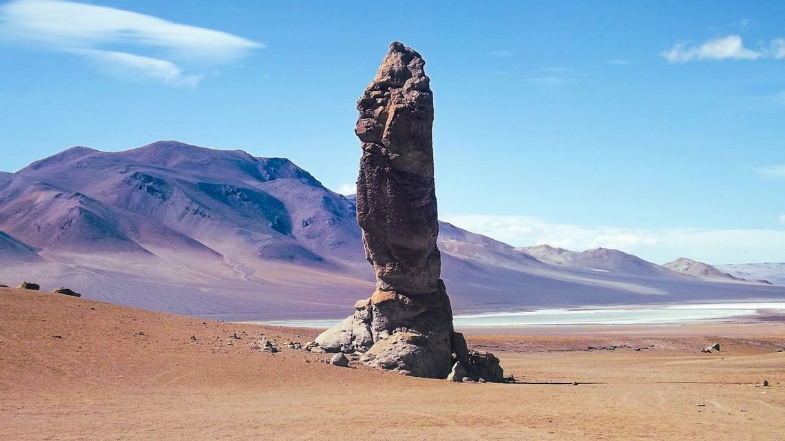 Una guglia di roccia imponente si erge in un vasto deserto, con montagne viola in lontananza e una distesa salina sotto un cielo azzurro e limpido.