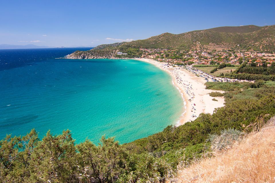 Una vista aérea de una playa de arena concurrida y en forma de media luna con agua turquesa, un pueblo costero y colinas verdes bajo un cielo azul claro.