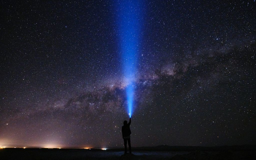 Una figura in silhouette punta il raggio blu di una torcia verso la Via Lattea in un cielo notturno stellato.