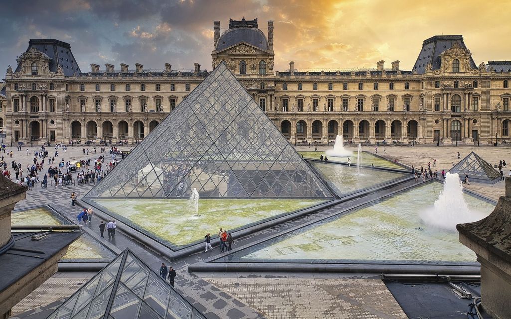 Una grande piramide di vetro e fontane circondate da persone in un cortile storico sotto un cielo caldo al tramonto.
