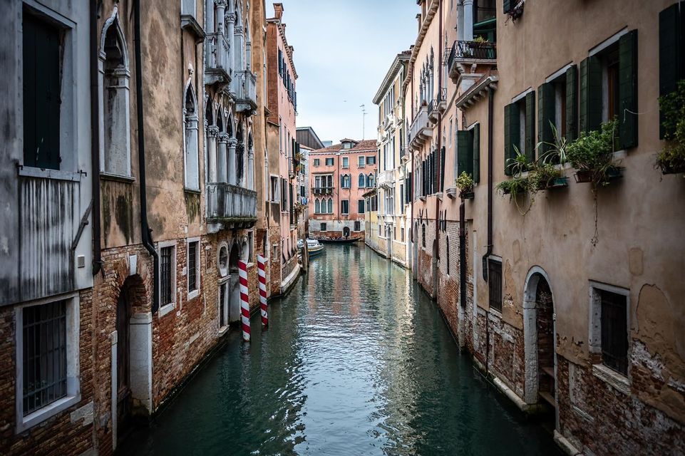 A narrow canal flows between tall, weathered buildings with balconies and shutters under an overcast sky.