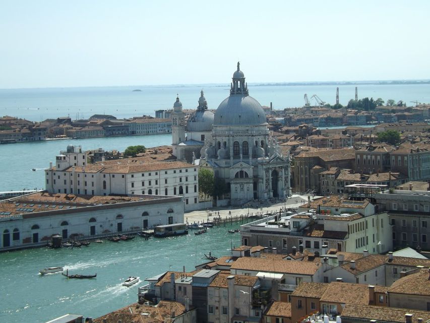 An aerial view of a city with a large domed basilica and terracotta-roofed buildings next to a canal with boats.