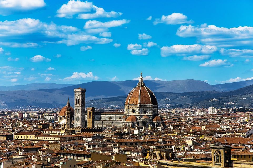The Florence Cathedral's dome and bell tower rise above the city's terracotta roofs, with mountains in the background under a blue sky.