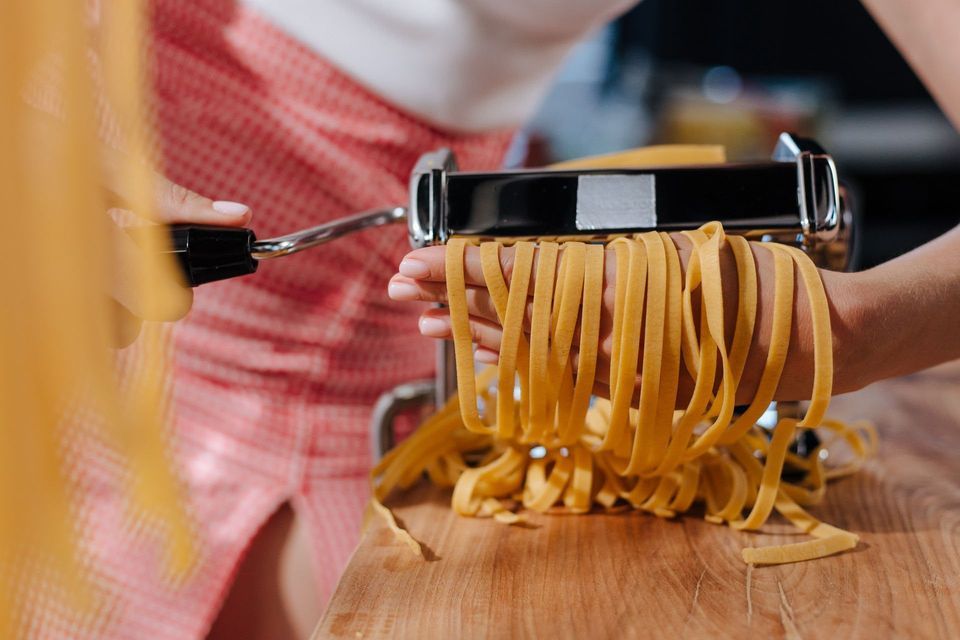 A person's hands catch strands of fresh fettuccine pasta coming out of a manual pasta maker over a wooden board.