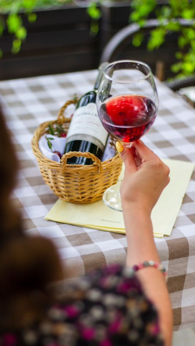 A person holds a glass of red wine over a checkered tablecloth with a wine bottle in a wicker basket.