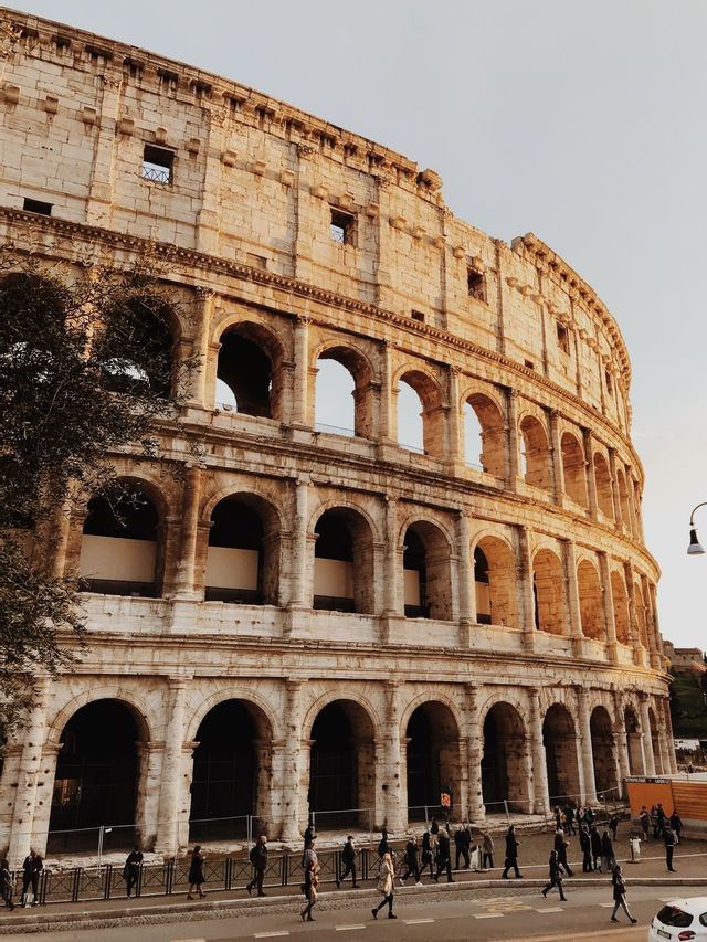 The exterior of a large ancient stone amphitheater with multiple tiers of arches, illuminated by the setting sun.