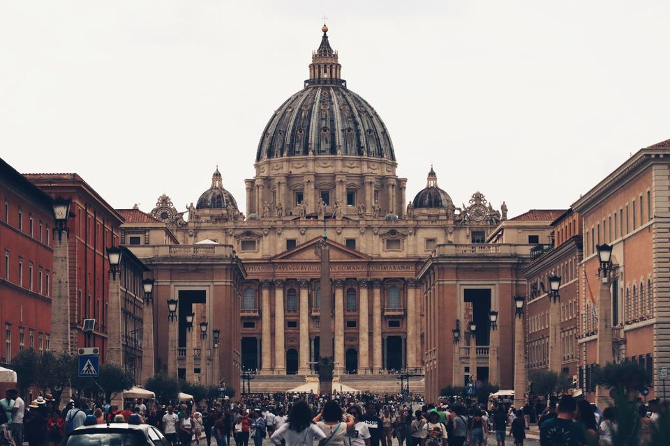 A large crowd gathers on a wide street leading to the grand facade and dome of a historic basilica under an overcast sky.
