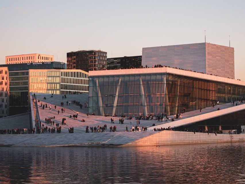 People walking and gathering on the sloping marble roof of a modern waterfront opera building at sunset.