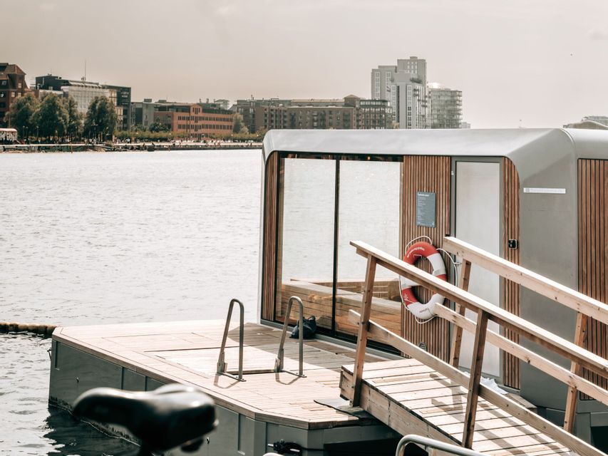 A modern floating wooden sauna with large glass windows and a lifebuoy rests on the water near an urban shoreline.