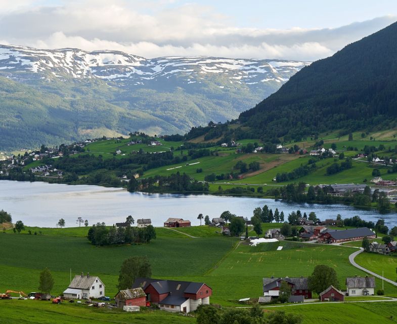 Rural houses and green farm fields along a lake with snow-capped mountains in the background.