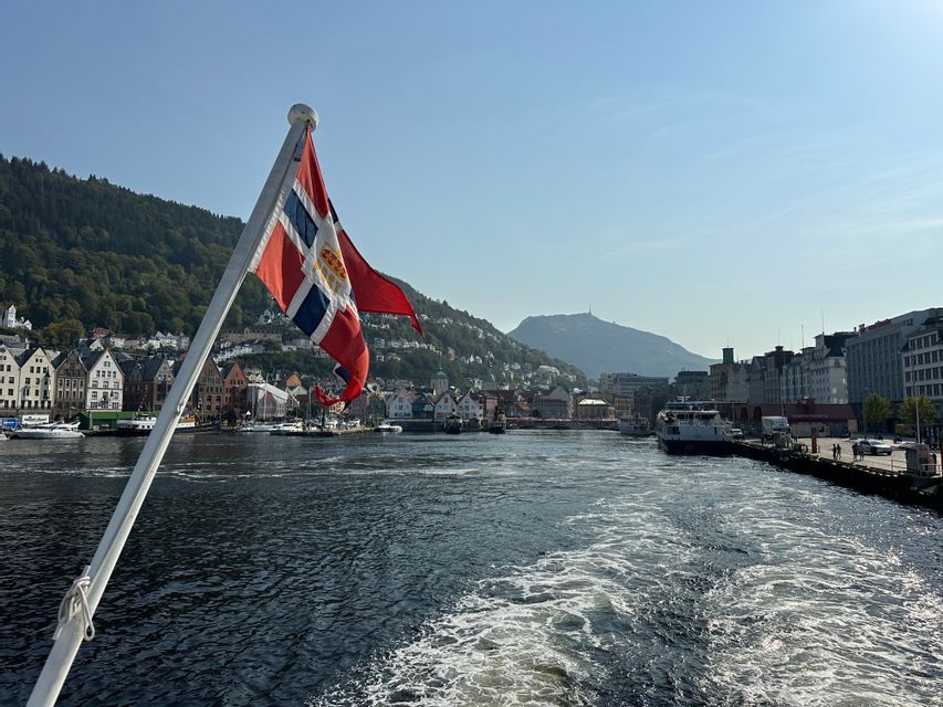 A boat leaves a waterfront harbor lined with historic buildings and green hills under a clear blue sky.