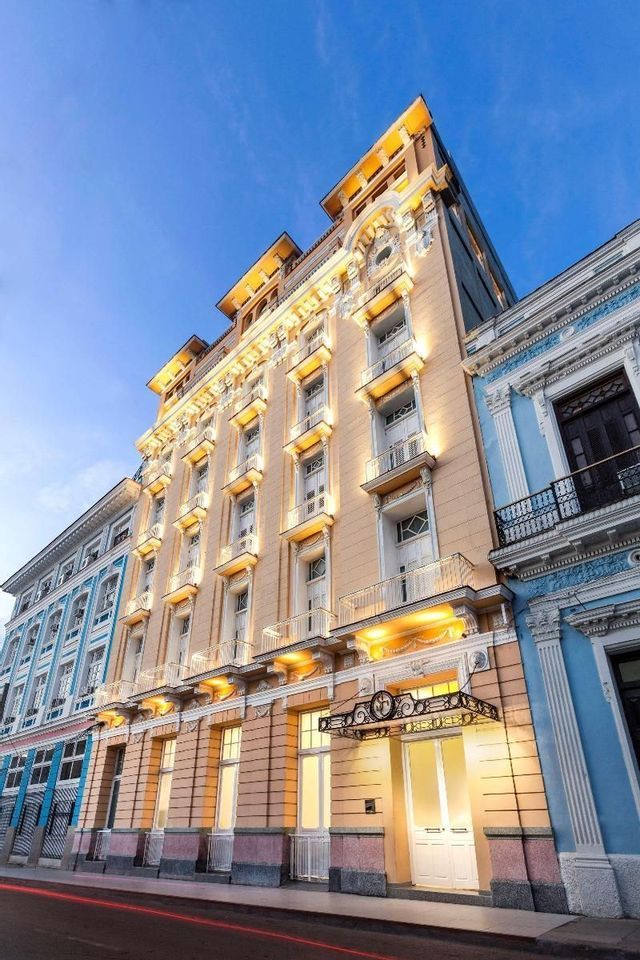 The ornate facade of a tall, beige hotel is illuminated by warm lights against a deep blue sky at dusk.