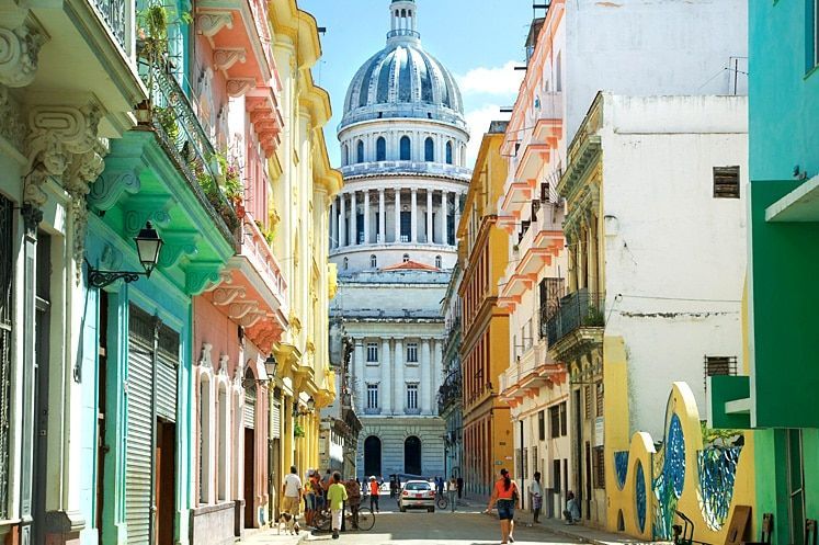 A city street lined with colorful buildings leads towards a large, classical-style domed capitol building under a clear sky.