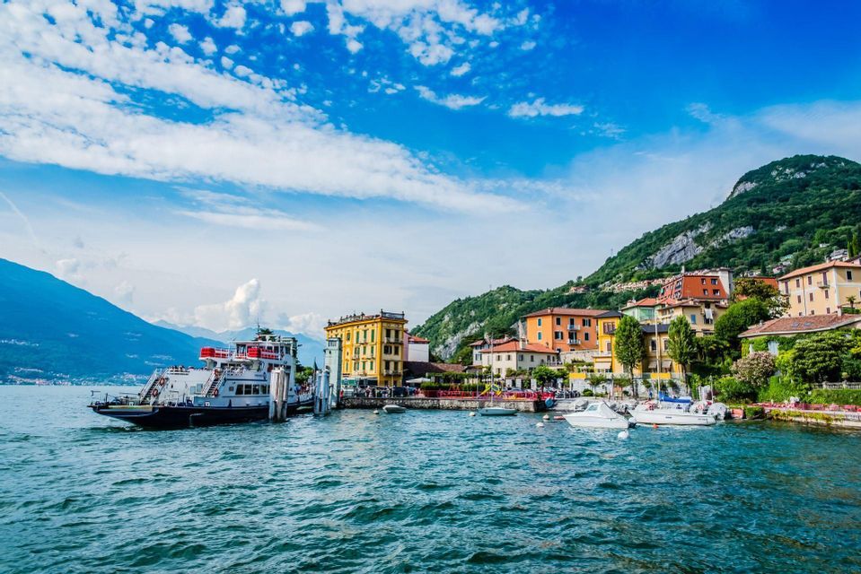 A ferry boat is docked at a pier in a colorful lakeside town, with lush green mountains rising behind it under a blue, cloudy sky.