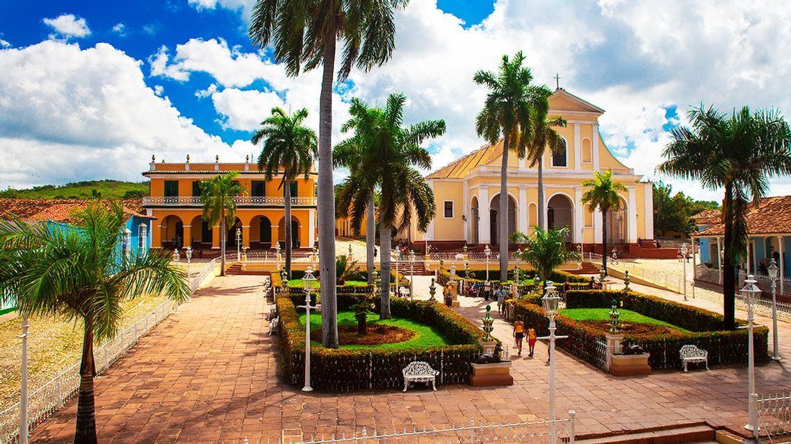 A sunny town square featuring yellow colonial buildings, tall palm trees, and a central garden under a bright blue sky with clouds.
