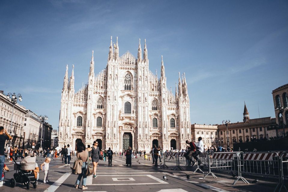An ornate, white Gothic cathedral stands behind a bustling public square filled with people on a sunny day.