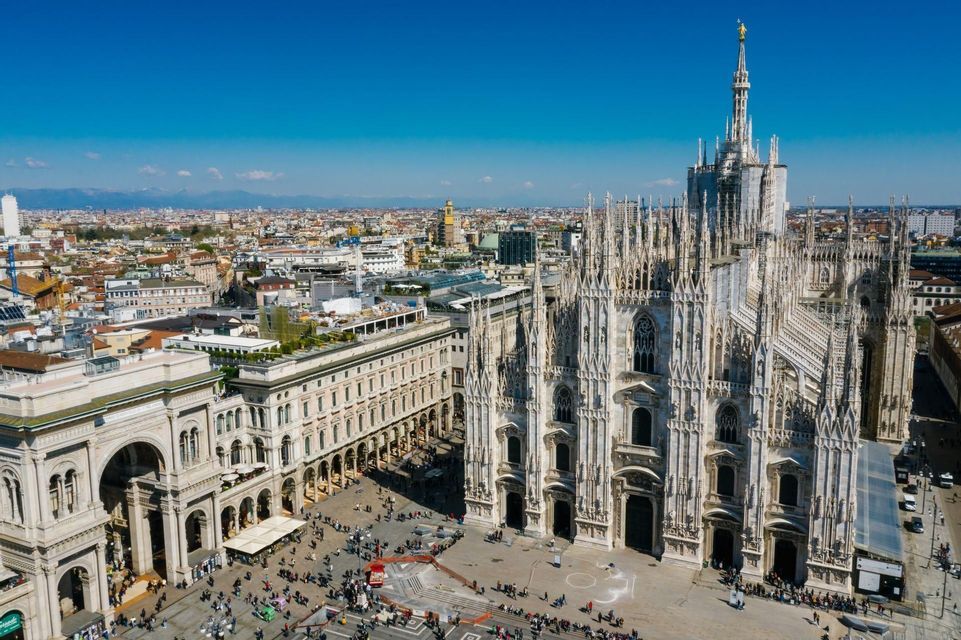 High-angle view of a large, ornate Gothic cathedral and a busy city square with crowds of people under a bright blue sky.