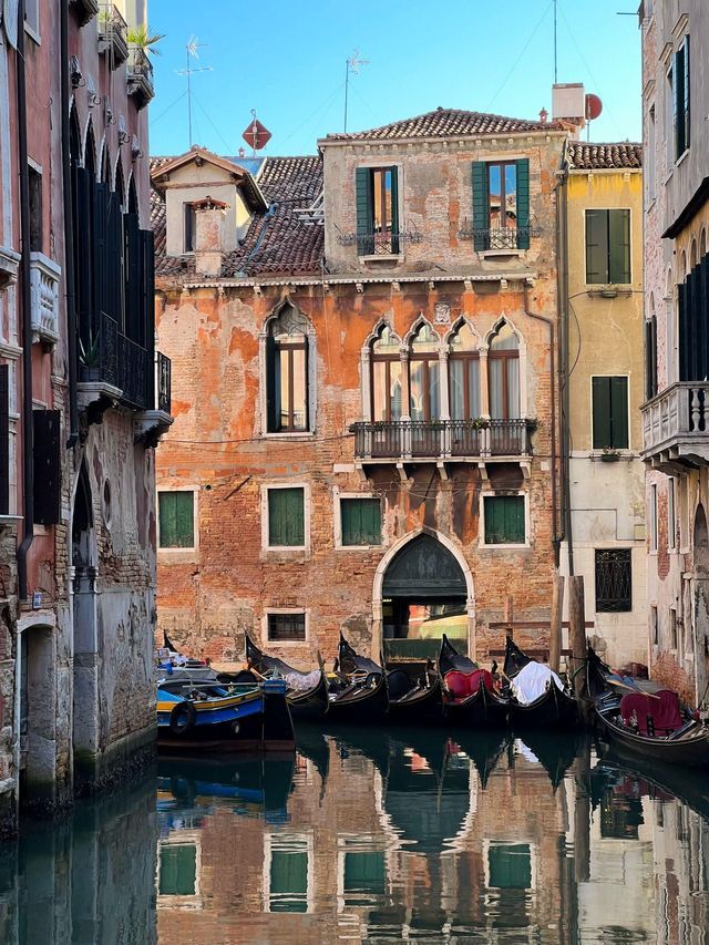 Gondolas are docked in a narrow canal, with their reflections mirrored in the water beside weathered brick buildings.