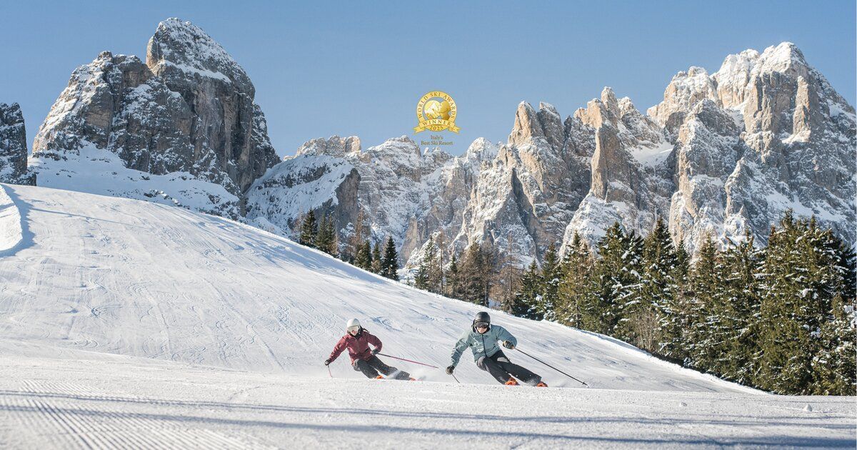 Due persone sciano su un'ampia pista battuta, con uno sfondo di montagne innevate sotto un cielo azzurro e limpido.