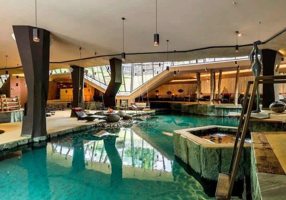 L'interno di una spa moderna con una grande piscina turchese, altalene sospese, aree lounge e grandi pilastri strutturali.