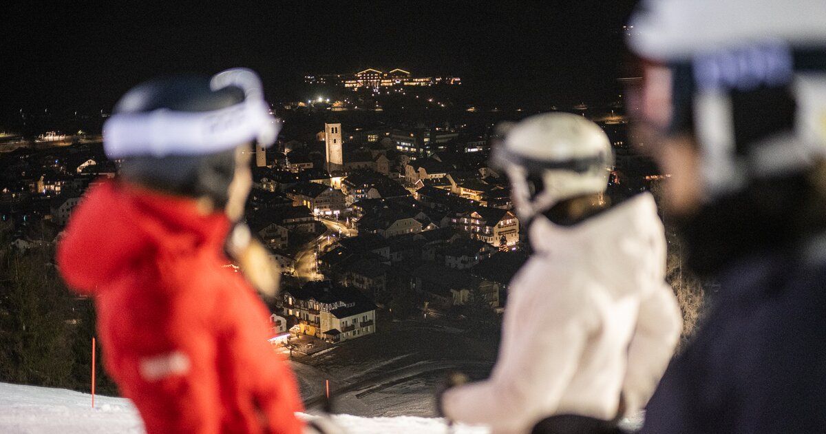 Un viaggio di gruppo WeRoad di sciatori su una pista di notte, che guarda giù un villaggio di montagna sottostante illuminato a festa.