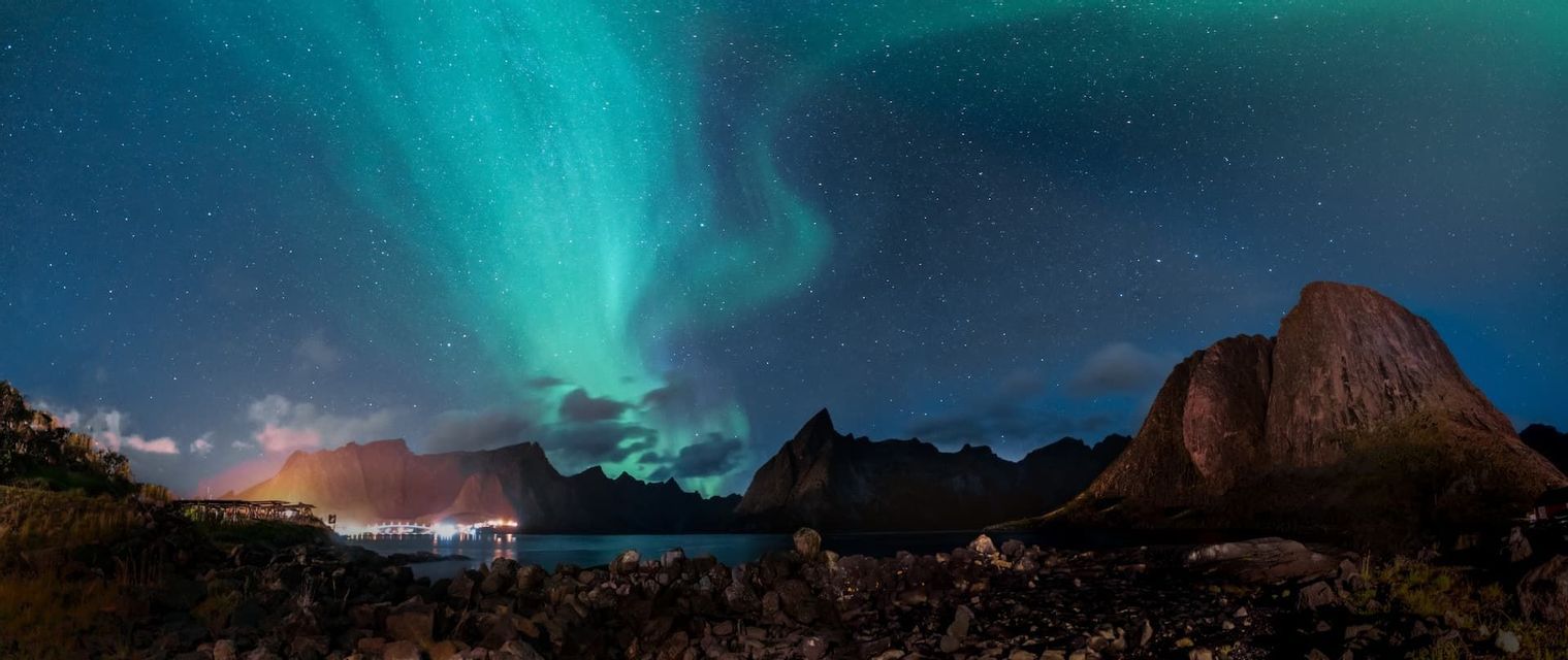 Das grüne Nordlicht leuchtet an einem sternklaren Nachthimmel über einem Fjord, mit Bergsilhouetten und einer felsigen Küste darunter.