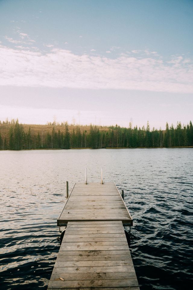 Un ponton en bois avec une petite échelle s'avance dans un lac, une forêt dense bordant la rive lointaine sous un ciel partiellement nuageux.