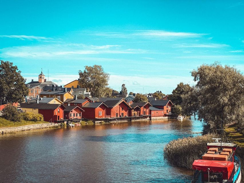Une rangée de maisons en bois rouges borde la rive d'une rivière sous un ciel bleu clair, avec des arbres en arrière-plan.
