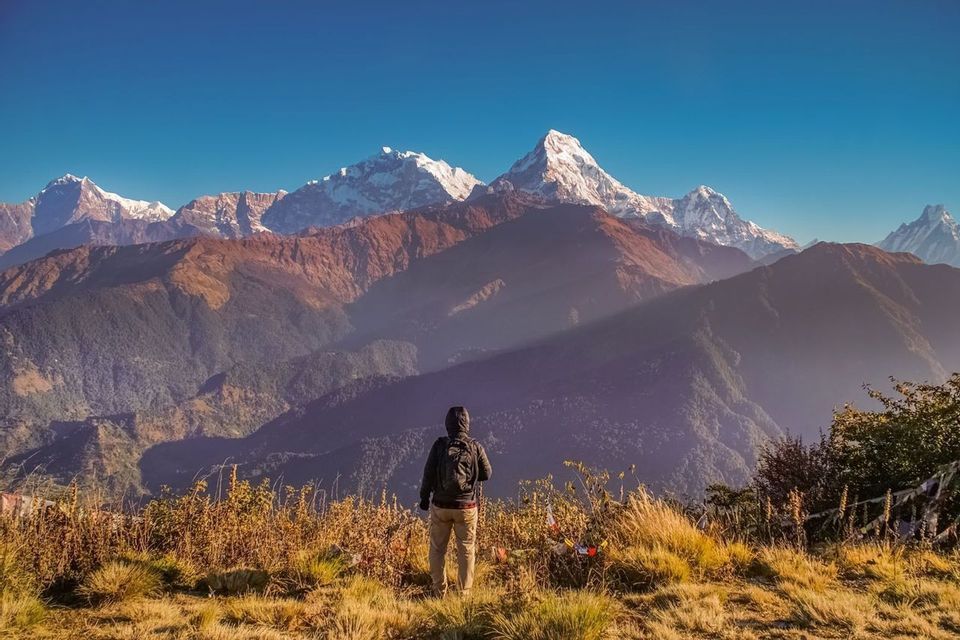 Un escursionista con uno zaino si trova su un crinale erboso, osservando una vasta catena di montagne innevate sotto un cielo azzurro e limpido.
