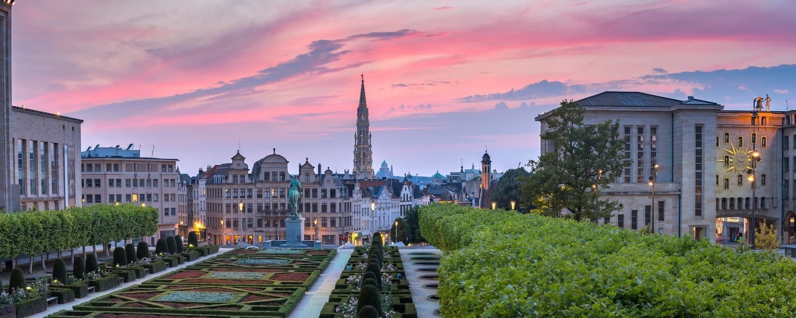Ein Panoramablick über einen formalen Garten auf eine historische europäische Stadtsilhouette mit einem hohen Kirchturm unter einem leuchtend pinkfarbenen Sonnenuntergang.