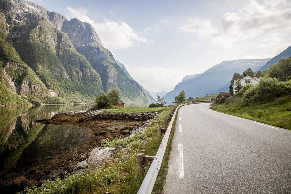 A paved road winds along the edge of a fjord, with steep, green mountains rising from the water under a partly cloudy sky.