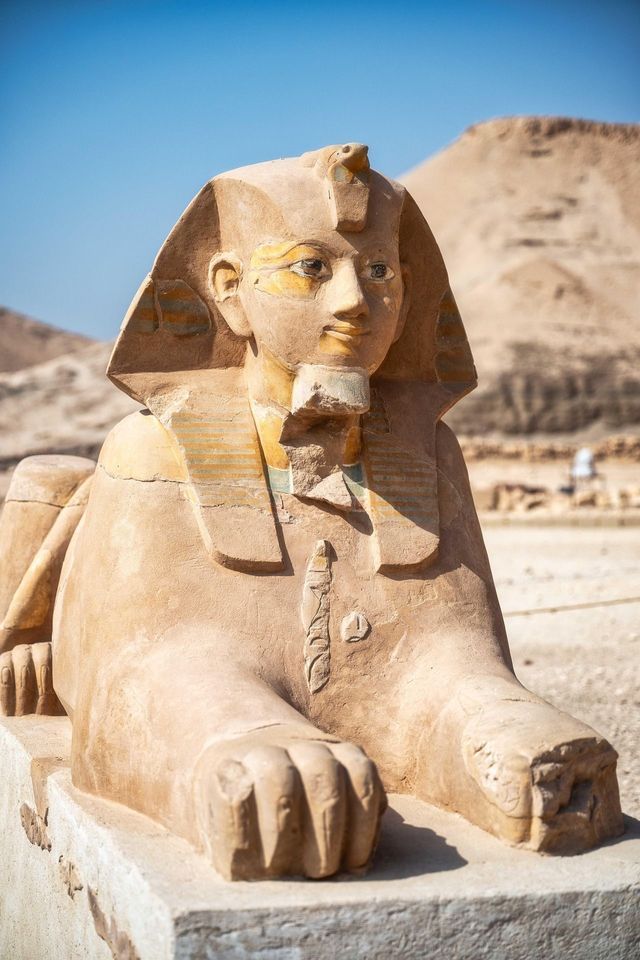 Eine antike Sandstein-Sphinx-Statue ruht auf einem Sockel bei hellem Tageslicht in einer Wüstenlandschaft.