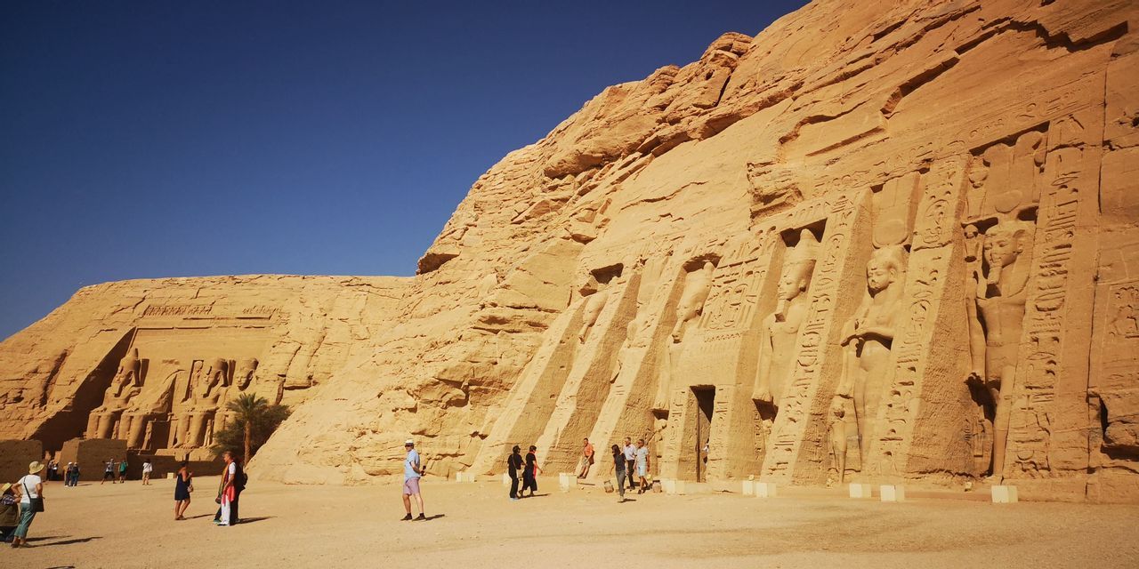 Touristen gehen auf sandigem Boden vor kolossalen antiken Statuen, die in eine Sandsteinklippe gehauen sind, unter einem klaren blauen Himmel.