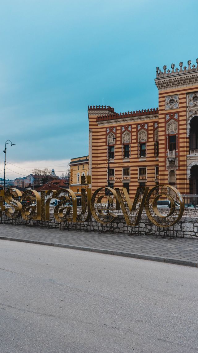 Un grande cartello dorato con la scritta 'Sarajevo' su un marciapiede di fronte a un edificio ornato e a strisce sotto un cielo nuvoloso.
