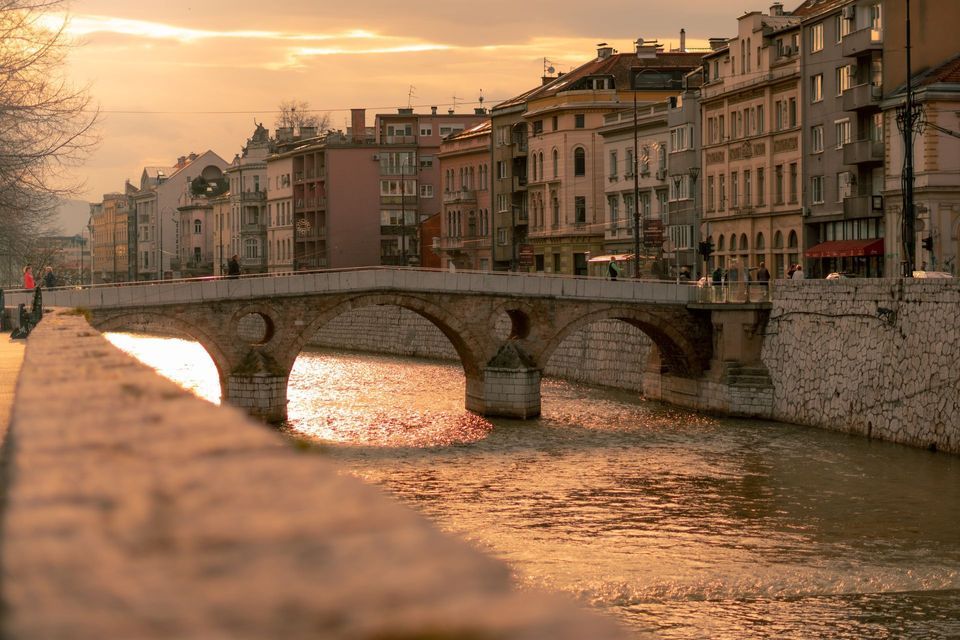 Un ponte ad arco in pietra attraversa un fiume in una città, con l'acqua che riflette la luce dorata del tramonto.
