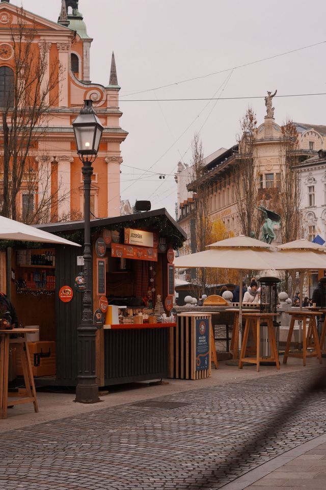 An outdoor market stall and seating area on a cobblestone street in a historic city square under an overcast sky.