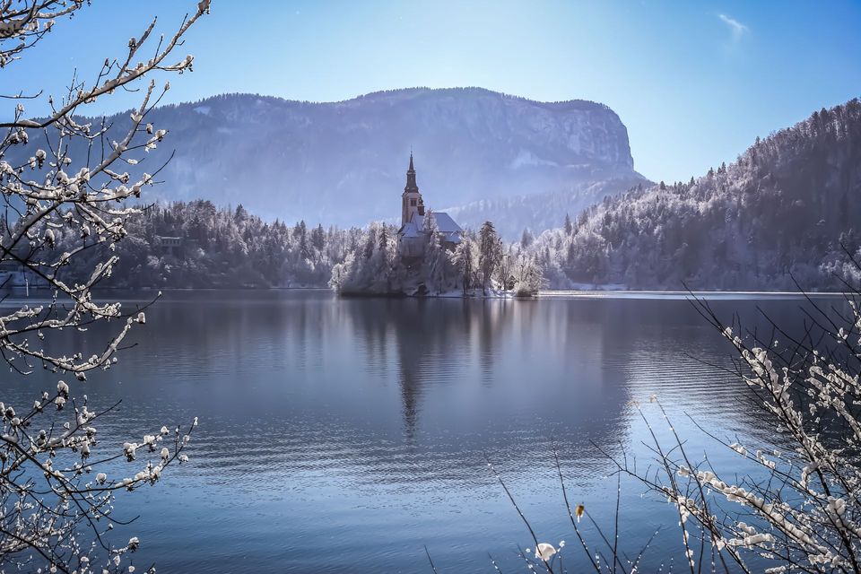 A church on a snow-covered island in a lake, surrounded by frosty trees and distant mountains under a clear blue sky.