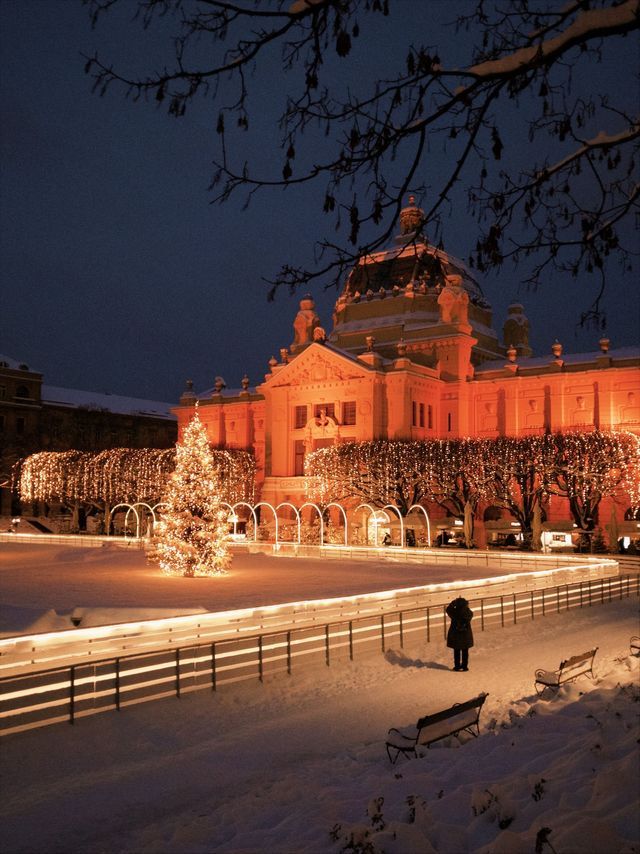 An illuminated building overlooks a snowy outdoor ice rink with a large Christmas tree at night.