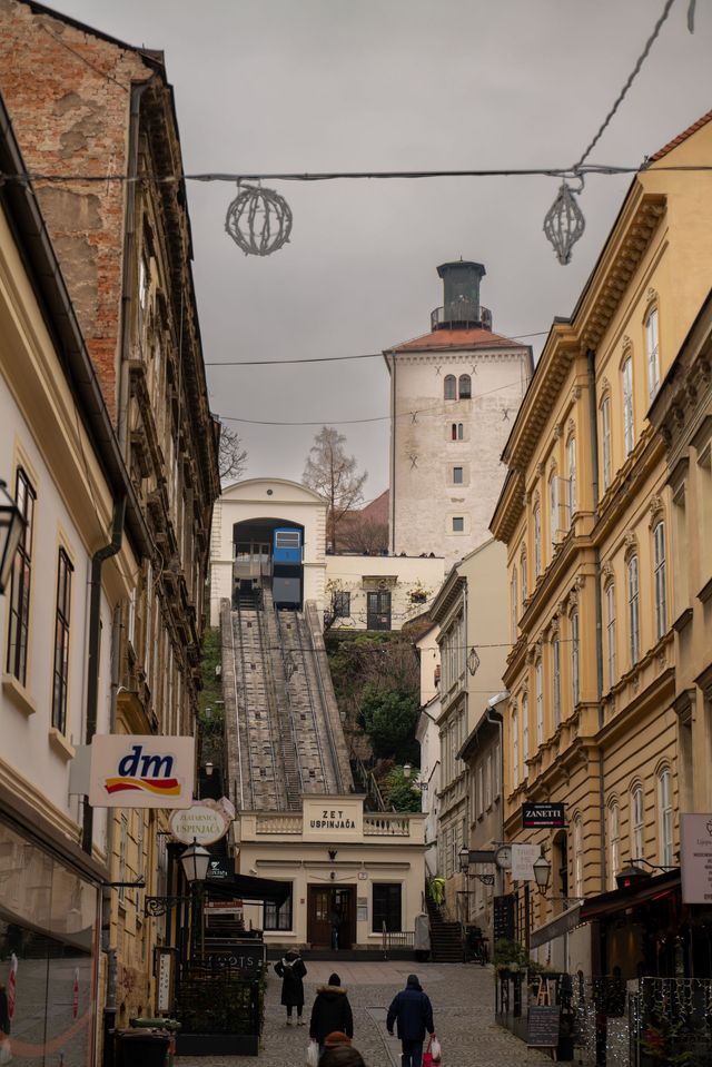 A blue funicular climbs a steep track on a narrow city street, with a historic tower visible in the background.