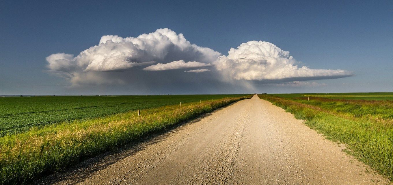 Una lunga strada sterrata si estende attraverso ampi campi verdi sotto una grande nuvola temporalesca bianca contro un cielo azzurro.