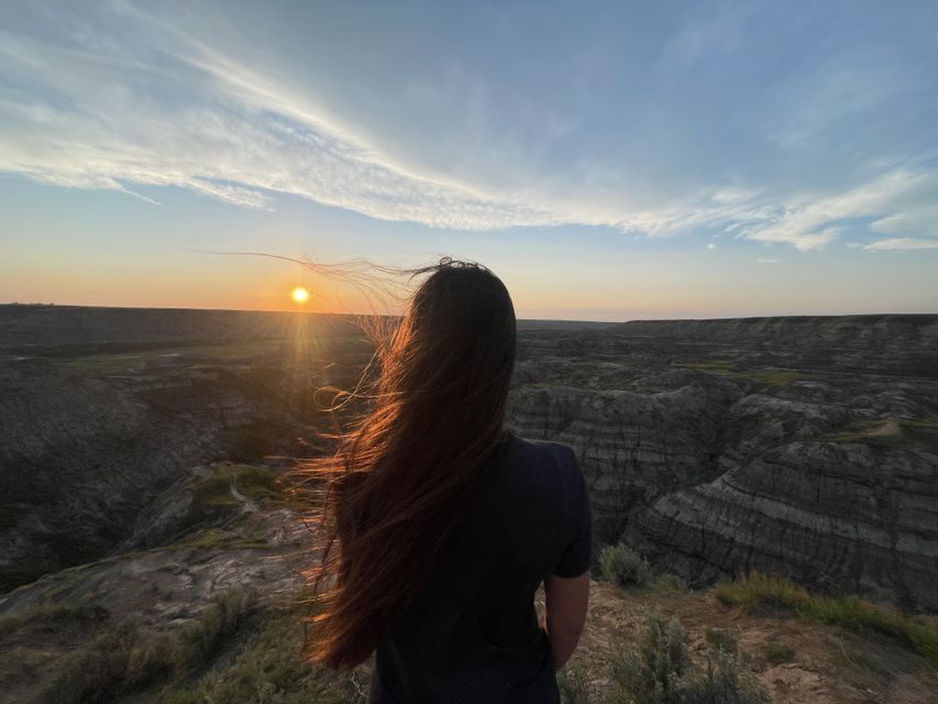 Una donna vista di spalle, con lunghi capelli al vento, ammira un vasto paesaggio di canyon al tramonto.