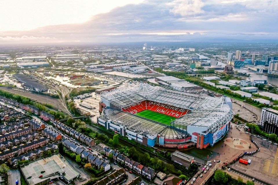 Una veduta aerea di un grande stadio di calcio con spalti rossi e un campo verde circondato da un paesaggio urbano.