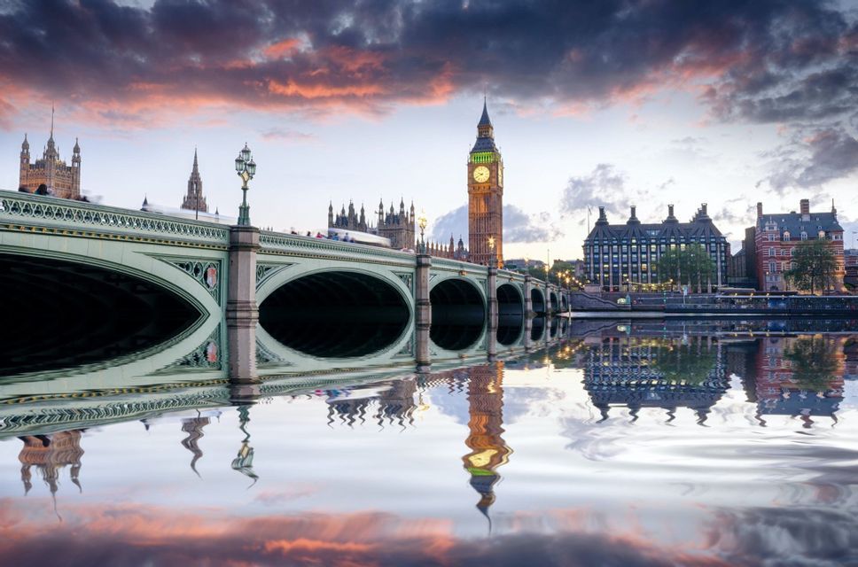Un ponte ad arco attraversa un fiume verso una torre dell'orologio illuminata, con nuvole colorate del tramonto che si riflettono sull'acqua.
