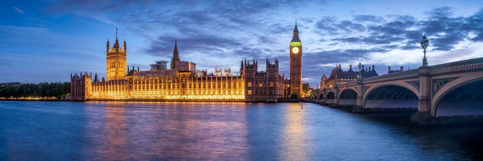 Un parlamento gotico illuminato e una torre dell'orologio accanto a un ampio fiume al crepuscolo con un ponte ad arco
